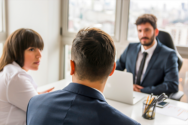 A person in a business suit is seen from behind, sitting and facing two professionals across a table, possibly discussing workers' compensation legal matters.